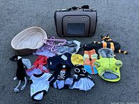 Full view of dog bed, carrier bag, assorted dog clothes, and toys arranged on pavement.