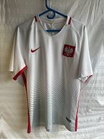 Front view of white Poland 2018-19 home football shirt with red Nike logo and Poland national team emblem, hanging on blue hanger.
