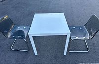 White square table and two transparent plastic chairs with metal cantilever bases placed on outdoor asphalt