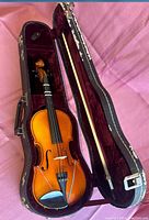 Half size violin with bow inside a worn black hard case lined in maroon velvet, photographed from above.