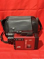 JVC GR-C7U camcorder in red with black focusing lens and attached viewfinder, positioned in front of black carry bag on red fabric background.