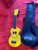 Yellow Mahalo U-Smile soprano ukulele next to black Mahalo branded carrying case on red background showing full body and headstock details.