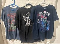 Three Marvel Comics t-shirts hanging on hangers showing front graphic design of Spider-Man, Fantastic Four, and Guardians of the Galaxy.