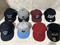 Front view of eight men’s snapback hats arranged in two rows of four on a patterned fabric background showing all embroidered logos clearly