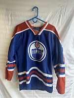 Front view of Edmonton Oilers Ryan Nugent-Hopkins hockey jersey hanging on a blue plastic hanger against a white backdrop.