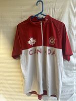 Front view of the Canada Olympic team jersey shirt, youth size XXL, hanging on a blue hanger, showing red top with white bottom, 'CANADA' text, maple leaf 04 emblem, and Olympic team logo.