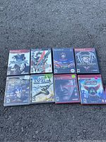 Photo of 8 game cases for PlayStation 2 and 3, arranged in two rows on asphalt surface.