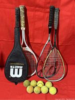 Four tennis racquets with various grips and one black Wilson Manta cover with eight used tennis balls on a red background.
