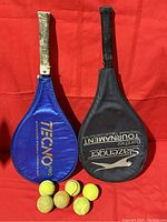 Top view of two tennis racquet covers, one blue Tecno Pro, one black Slazenger, with six tennis balls in front on red background.