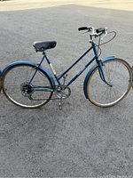 Blue vintage Raleigh bicycle shown on pavement, rear tire appears deflated, black saddle and grips, step-through frame design, metal fenders on front and rear wheels.