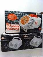 Two bubble machines in original packaging. White bubble machine white plastic body with orange bubble ring, black bubble machine with yellow bubble ring.