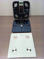 Two digital smart scales shown side by side on a surface, one black with LCD and sensors, one white with metal electrodes.