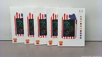 Front view of five sealed boxes of the LCD writing tablet, showcasing the dinosaur design and red-white striped border with an educational theme.