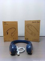 Blue neck fan device with USB charging cable placed in front of two cardboard boxes labeled R5 and S8 showing sketches of the neck fans.