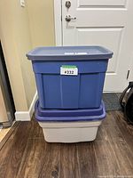 Two stacked plastic storage bins, a blue bin on top of a beige bin, shown closed with a lot number label.