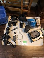 Photo showing a collection of three Canon cameras with multiple lenses and accessories on a white cloth, including a Canon EOS film camera, lenses, remote shutter release, battery grips, and boxes.