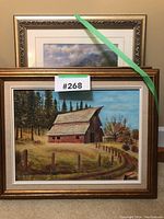 Photo showing two framed paintings stacked, front one depicting a red barn in a fall landscape with fence and trees, back one a mountain landscape print