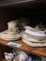 Shelf holding stacked cups, plates, and serving dishes including a gravy boat and a covered sugar bowl with floral pattern.