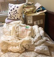 Assorted linens, blankets, quilts, and a decorative pillow arranged on a mattress with a window in background. Includes folded linens and quilts, some with lace and floral embroidery.