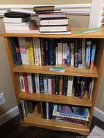 Photo showing a bookshelf with multiple books on two shelves and several books stacked on top, mostly hardcover and paperback novels and reference books.