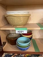 Bowls arranged in a cabinet shelf showing large mustard yellow and white mixing bowls on upper shelf, and smaller stacked colorful bowls with a medium brown bowl on the lower shelf.