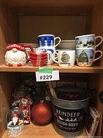 Shelf displaying Christmas mugs featuring snowmen, Christmas trees, and winter scenes, along with a red and clear glass reindeer figurine, metal bucket with reindeer text, orange glitter ball ornament, and a Santa snow globe.
