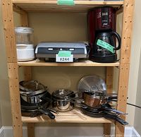 Shelf containing multiple kitchen items including a white Farberware Handy Steamer appliance, a silver vacuum sealer, an Oster red coffee maker without lid, and a collection of pots and pans with handles and lids