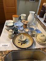 Photo showing the entire assortment of assorted fine china, pottery, and glassware arranged on a kitchen counter with detailed view of shapes and patterns visible.