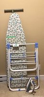 Full view of ironing board with floral cover, drying rack in front, and iron on floor.