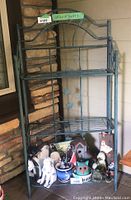 Front view of metal garden shelf showing decorative leaf designs and three wire shelves. Contents on shelf not included.