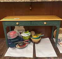 Front angle of oak top and green wooden frame console table with two drawers and round wooden knobs; includes kitchen items underneath but not included.
