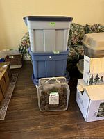 Stack of three Rubbermaid storage bins in blue, gray, and white colors with lids, placed on a wooden floor against a floral couch background.