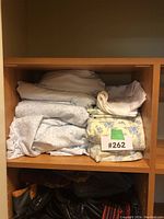 Multiple queen bed linens folded and stacked on wooden shelf, showing different colors and patterns including gray, white, and floral prints.