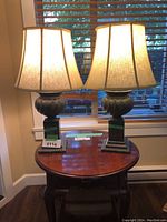 Pair of dark grey lamps lit on a wooden table in front of window blinds.