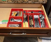 Drawer containing red trays with assorted kitchen cutlery and utensils including forks, knives, spoons, measuring spoons and other kitchen gadgets.