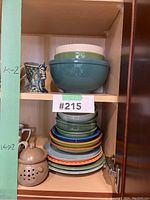 Shelf with various ceramic kitchenware including three mixing bowls stacked and several plates below plus a decorative ceramic lidded pot and a large detailed blue mug.