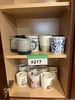 Two shelves of ceramic mugs showing assorted patterns and animal illustrations. Chip noted on one mug with green tape.
