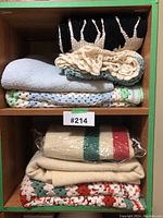 Lot of eight blankets displayed on two wooden shelves, showing different materials and patterns including knit, crochet, and wool Hudson's Bay style blanket.