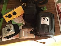 All five cameras arranged on a table, including three camera cases visible. Yellow Olympus waterproof camera stands out; two silver cameras (Minolta and HP); black Canon and cases.