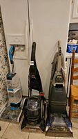 Front view of three carpet cleaners side by side on tile floor in kitchen area, showing overall used condition and model shapes.