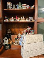Three shelves filled with a variety of Christmas-themed music boxes, ceramic figurines including angels, trees, and nativity scenes, along with visible stacked Mark Roberts boxes.
