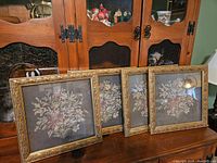 Set of four identical framed floral cross stitch pictures with ornate gold leaf-patterned frames.