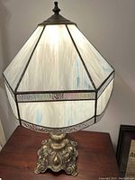 Full view of the lamp from the front, showing the stained glass shade illuminated and the ornate brass colored metal base.