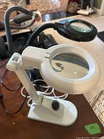 Both magnifying lights shown on a table. White magnifying light with adjustable arm and round lenses.