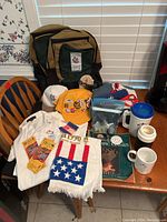 Full view of the lot on table showing backpacks, hats, shirts, towel, pins, cups, and books