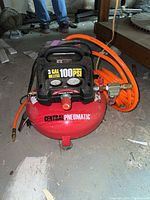 Full view of red Central Pneumatic pancake compressor with orange hose coiled beside it.