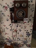 Replica vintage style wall phone mounted on floral wallpaper showing rotary dial, twin metal bells, wooden case, and handset with cord hanging.