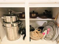 View of inside kitchen cabinet displaying stacked stainless steel pots, glass lids, amber glass cookware, and burner covers.