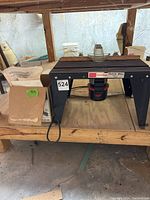 Photo showing Sears Craftsman router table with attached router and nearby router accessories on wooden shelving in a basement setting.