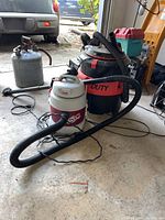 View of two shop vacuums, a large Kenmore Heavy Duty model in red and black with hose, and a smaller white and maroon Shop-Vac 7.0 amp vacuum with hose and power cord, all shown on garage floor.